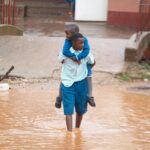 A man carries a child through a flooded city street during heavy rain, showcasing resilience.