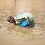 Young boy wading in a flood with a blue bowl collecting water. Outdoors scene.