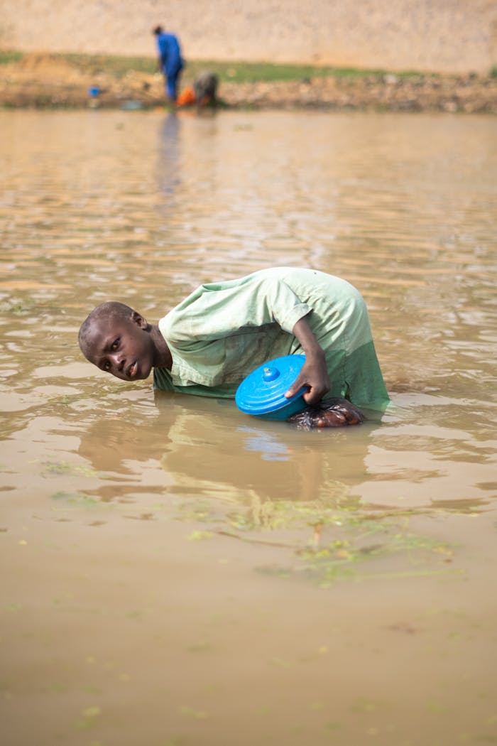 Young boy wading in a flood with a blue bowl collecting water. Outdoors scene.