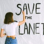 A woman in jeans and white top paints 'Save the Planet' on a wall, advocating environmental awareness.