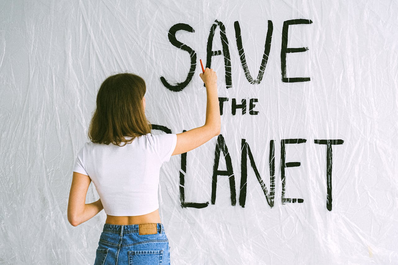 A woman in jeans and white top paints 'Save the Planet' on a wall, advocating environmental awareness.